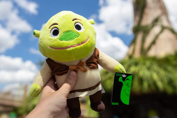 A hand holds up a Shrek plush toy in front of Shrek’s cottage in DreamWorks Land at Universal Studios Florida.