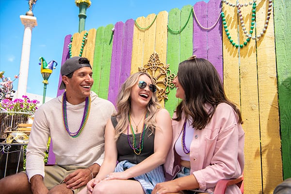 Three friends sit and laugh in front of a purple,yellow, and green fence decorated for Mardi Gras at Universal Studios Florida.