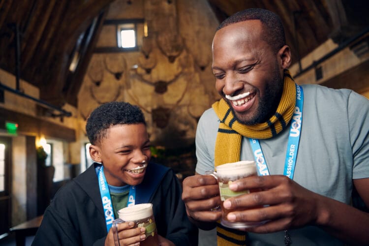 A father and son laughing while they drink Butterbeer™ with a line of frothy foam on their upper lips.