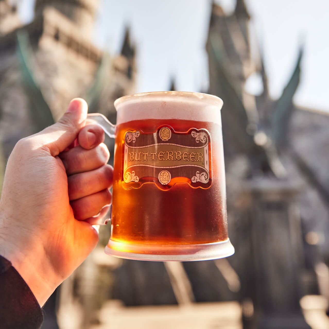 A person taking a photo of their mug of Butterbeer™ with Hogwarts™ castle in the background.