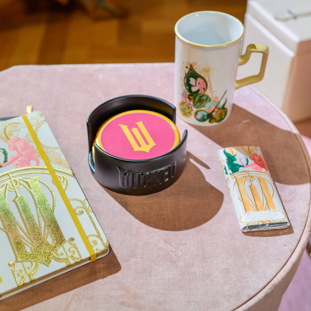 A pink desk topped with a Wicked themed notebook and cup, a stack of pink and yellow coasters with a W on the center, and a matcha chocolate bar.