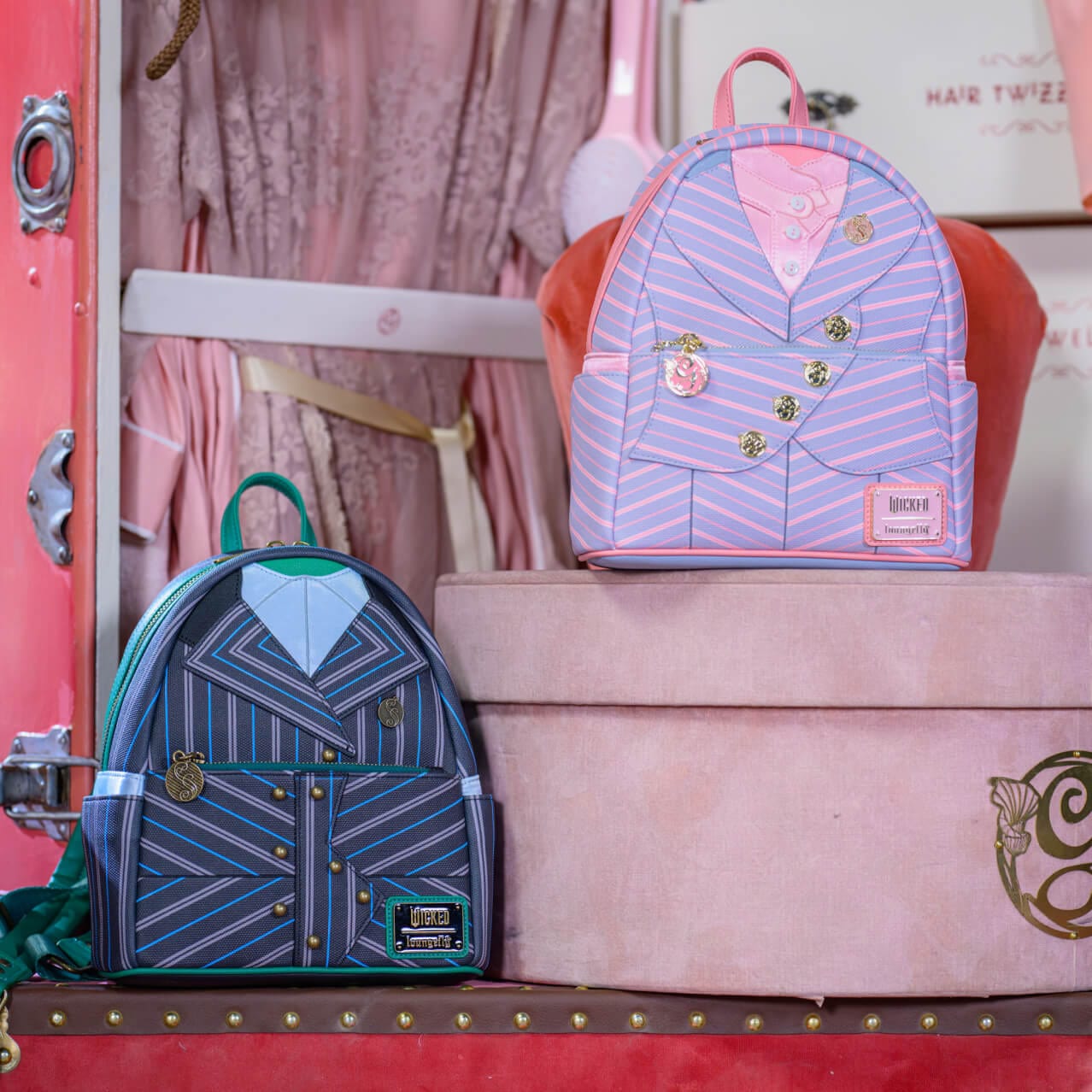 Two backpacks designed to look like Elphaba and Glinda's school uniforms sitting in front of a pink wardrobe.